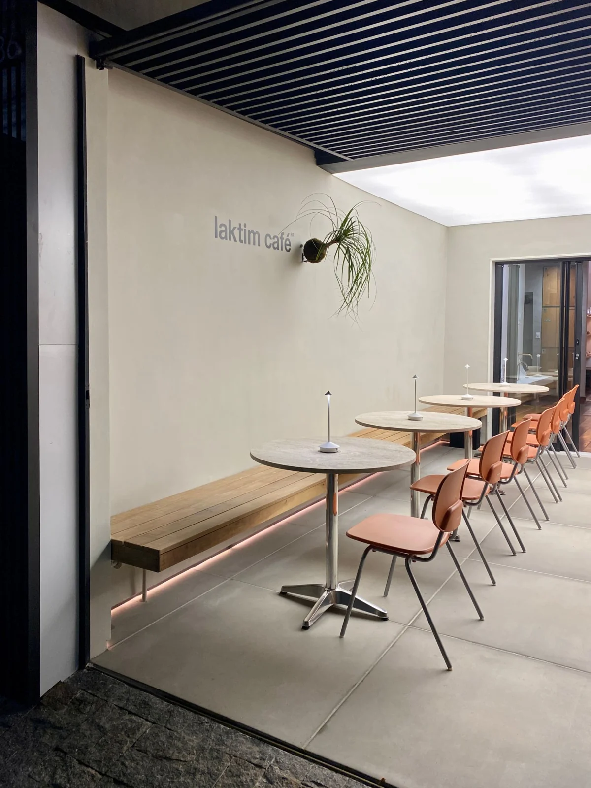 Laktim Café à noite com luz branca: banco de madeira, mesas de pedra, luminárias e cadeiras terracota sob o pergolado