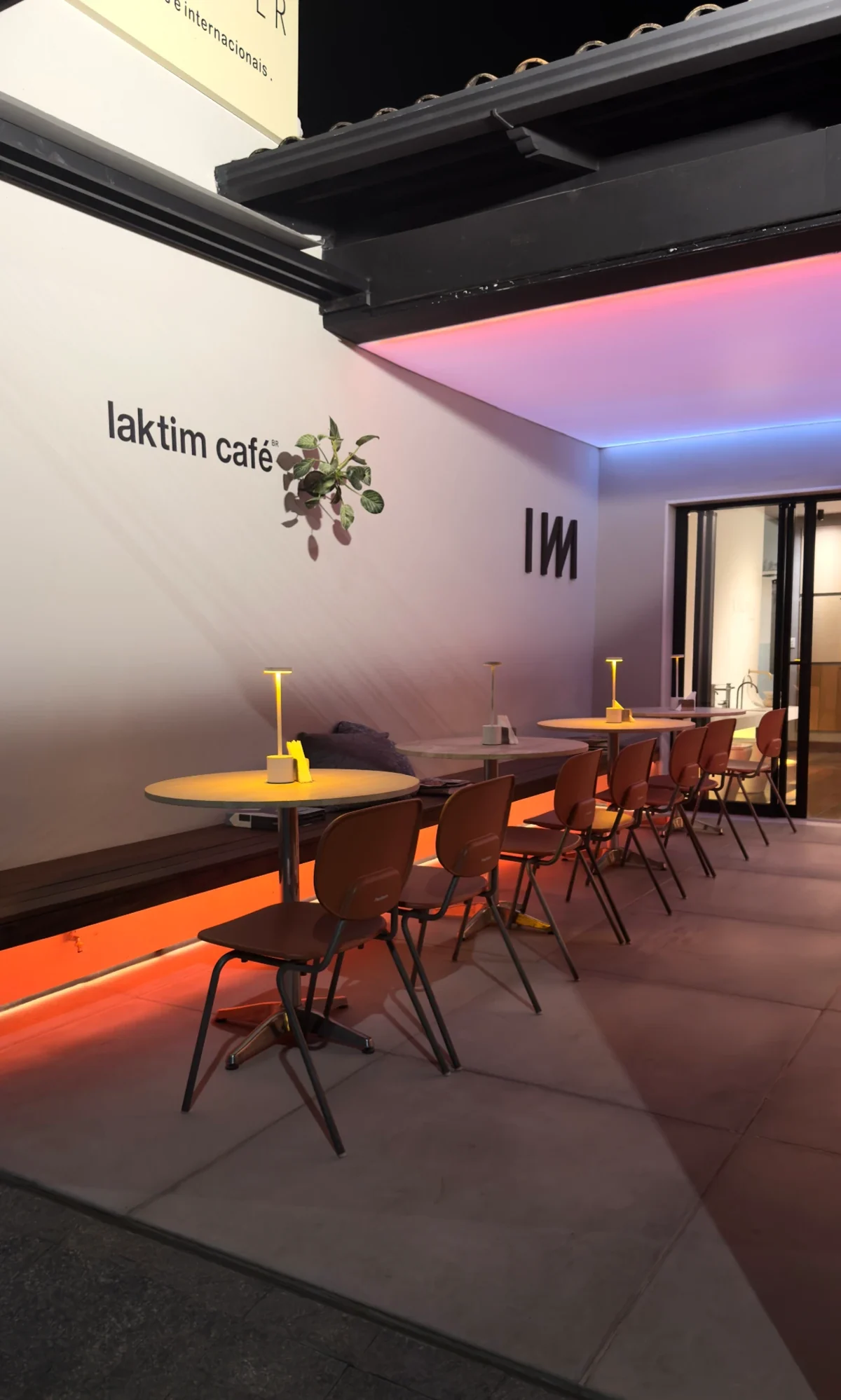 Laktim Café à noite: mesas com luminárias acesas, cadeiras terracota, o letreiro e o símbolo IИИ na parede