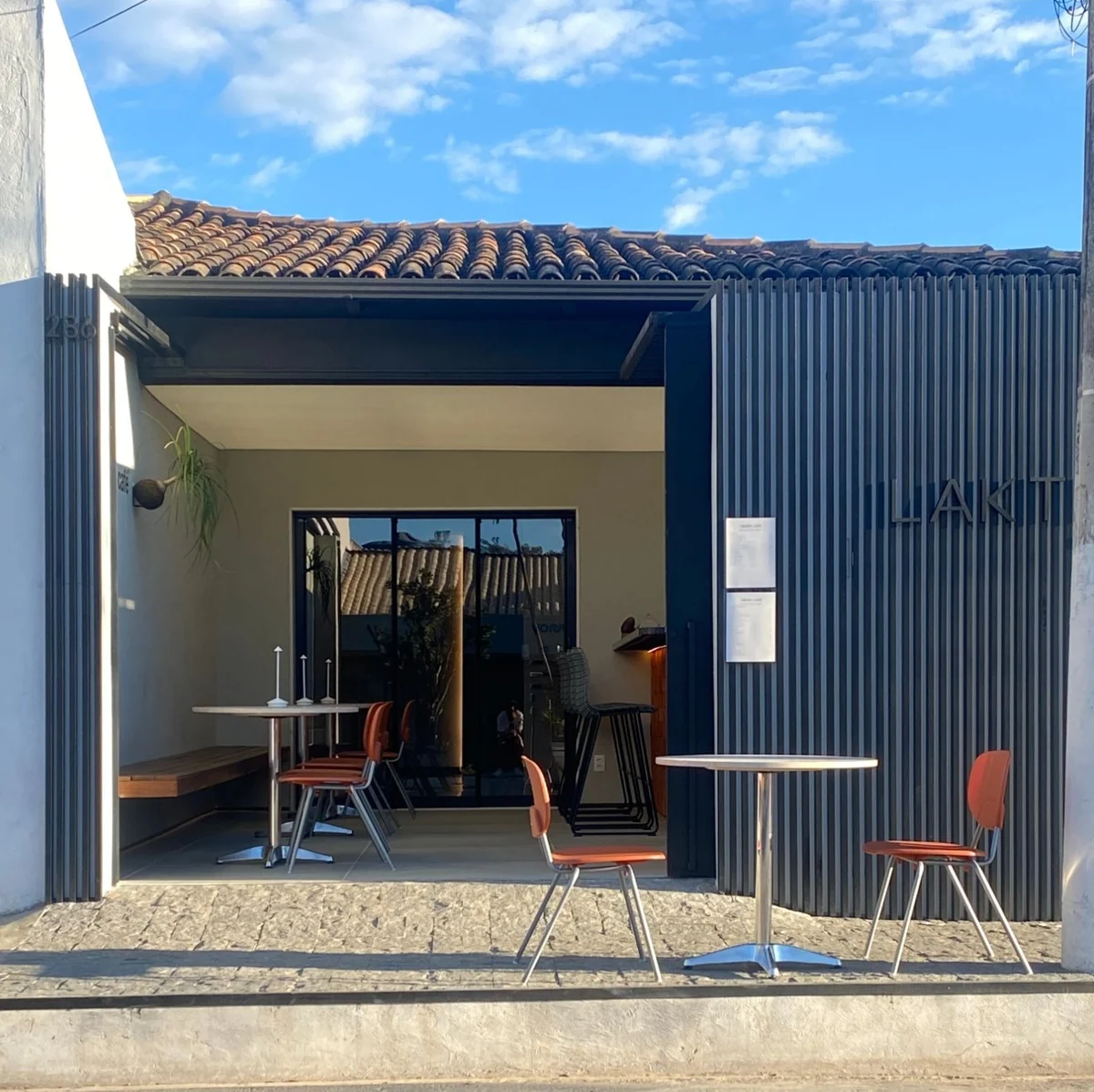 Fachada da Laktim Design de dia, com o Laktim Café aberto: mesas e cadeiras terracota na calçada e ripas pretas